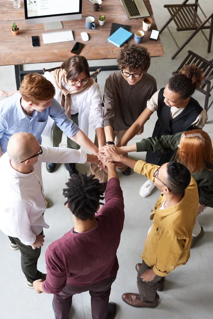 about-02 A diverse group of professionals joins hands in a team-building exercise indoors.